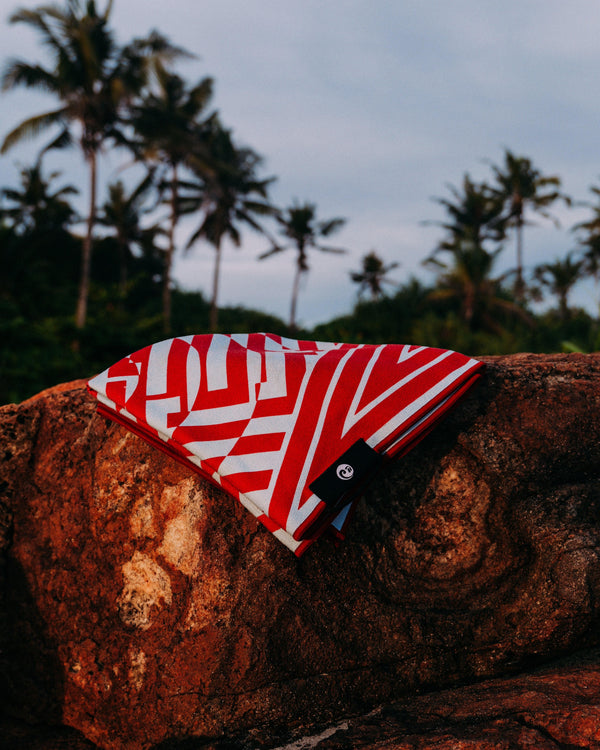 Tide and Trail Towel Ripple by Happy Faced Towels on a rock by the beach palm trees blow in the breeze behind