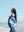 A woman wrapped in a colorful blue towel stands on the beach, gazing peacefully toward the horizon with a slight smile on her face. The towel features a bold, abstract design with pink and white elements. The ocean waves gently roll onto the shore behind her, and the sky is clear and bright, enhancing the serene and relaxed beach atmosphere. Her long dark hair flows slightly in the breeze, adding to the calm and carefree mood of the scene.