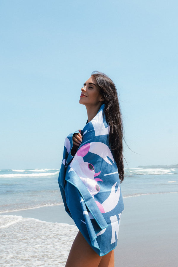 A woman wrapped in a colorful blue towel stands on the beach, gazing peacefully toward the horizon with a slight smile on her face. The towel features a bold, abstract design with pink and white elements. The ocean waves gently roll onto the shore behind her, and the sky is clear and bright, enhancing the serene and relaxed beach atmosphere. Her long dark hair flows slightly in the breeze, adding to the calm and carefree mood of the scene.