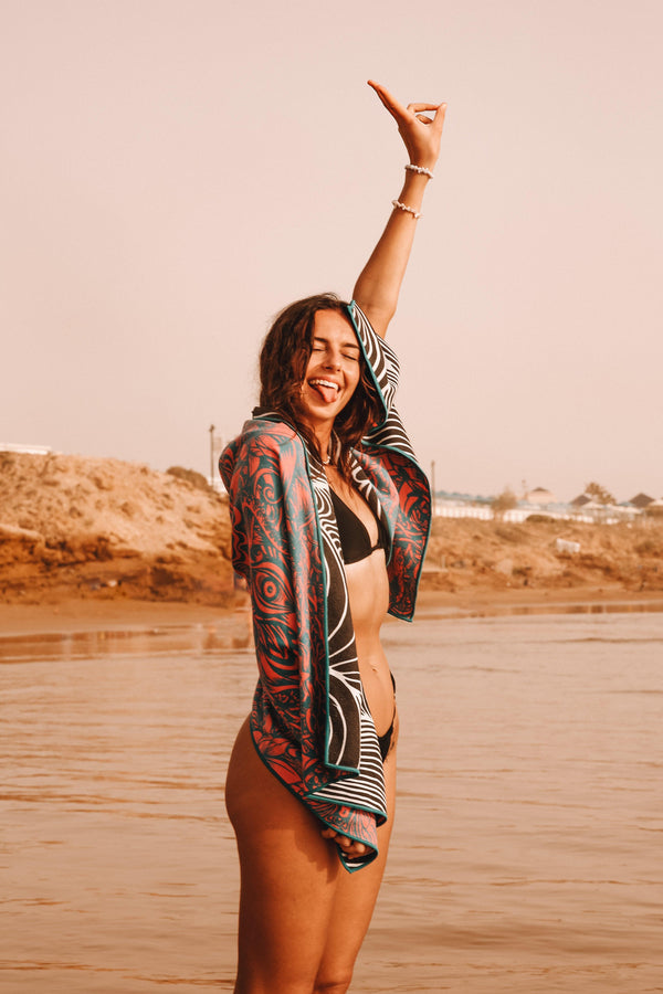 A woman draped in a Happy Faced Towel on the beach throws up a peace sign and a smile with her tongue out