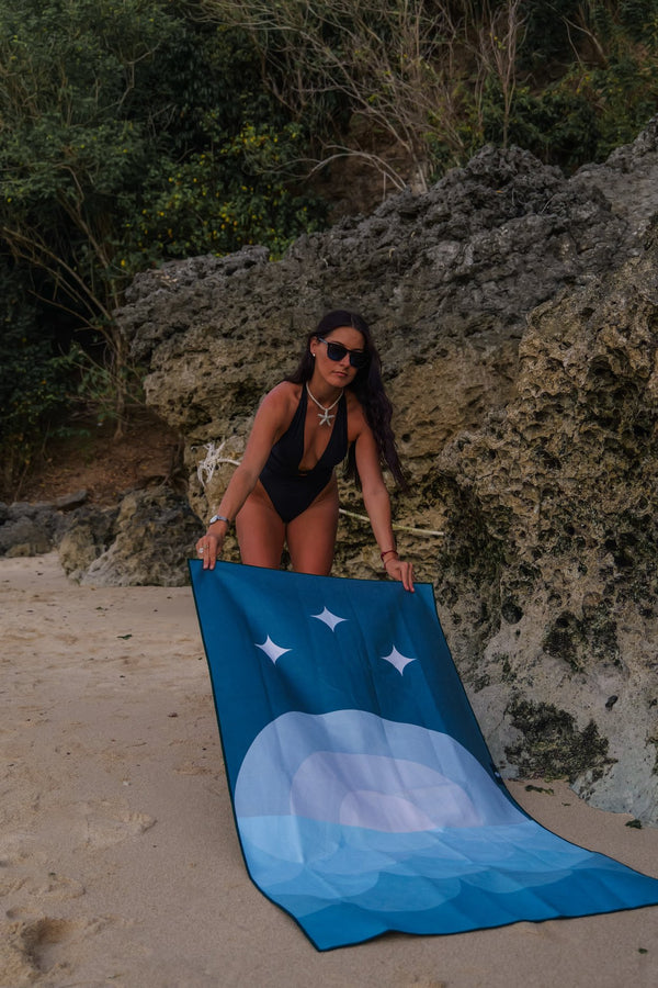 A woman wearing a black swimsuit and sunglasses is laying out a blue beach towel on the sand near a rocky shoreline. The towel features a minimalist design with abstract shapes and white star-like elements. The surrounding environment includes textured rock formations and greenery in the background, adding a natural and rugged feel to the scene. The woman, with a starfish necklace, appears focused as she prepares her space for relaxation, capturing a moment of calm at the beach.