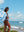 A woman in a white bikini stands on a rocky beach, holding up a large blue beach towel with a colorful abstract design and bold white text. The towel is caught in the breeze, billowing behind her. She is smiling and looking toward the ocean, with the bright blue sea and white-capped waves stretching into the horizon. The sky is clear with a few scattered clouds, creating a vibrant and peaceful beach atmosphere.