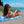 A woman in a white bikini lounges on a colorful beach towel with a vibrant, abstract design. She is reclining on the sandy beach, smiling and looking off to the side, with her hand resting on her head. Behind her, there are rocky outcrops and the bright blue ocean with small waves, along with a partly cloudy sky, creating a relaxed and sunny coastal scene.