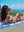 A woman in a white bikini lounges on a colorful beach towel with a vibrant, abstract design. She is reclining on the sandy beach, smiling and looking off to the side, with her hand resting on her head. Behind her, there are rocky outcrops and the bright blue ocean with small waves, along with a partly cloudy sky, creating a relaxed and sunny coastal scene.