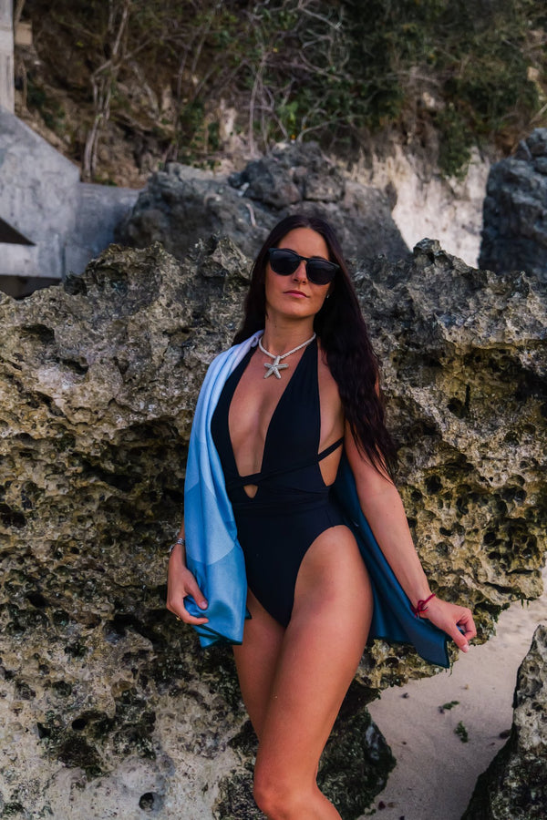 A woman wearing a black one-piece swimsuit and sunglasses stands confidently against a rocky backdrop at the beach. She has a light blue towel draped over one shoulder and wears a starfish-shaped necklace, adding a nautical touch to her look. The textured rocks behind her create a rugged, natural setting, while her pose and expression convey a sense of calm and strength. The combination of her attire and the scenery evokes a stylish, beachy atmosphere with a hint of adventure.