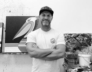 Artist Erik Abel standing in front of his painting of a pelican with arms crossed wearing baseball cap for Happy Faced Towels.