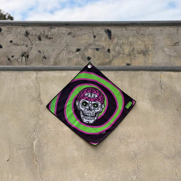 Happy Faced Attack Skateboarding Towel displayed on a concrete skatepark ledge. The artist-designed microfiber skate towel features a detailed skull with an exposed brain surrounded by hypnotic green and purple spiral rings, creating a bold horror-inspired graphic influenced by classic skateboarding art.