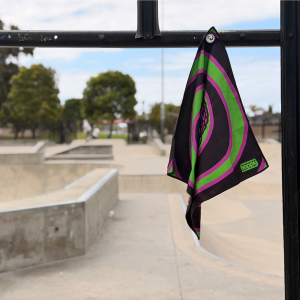 Happy Faced Attack Skateboarding Towel magnetically attached to a metal skatepark fence. The microfiber skate towel showcases a skull with an exposed brain surrounded by vivid green and purple spiral graphics, inspired by vintage horror and skate culture artwork. The built-in magnet keeps the towel securely attached and easily accessible while skating.