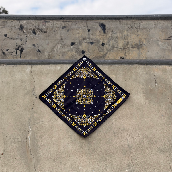 Happy Faced Bandito Skateboarding Towel displayed on a concrete skatepark. The artist-designed microfiber skate towel features a black bandana-inspired pattern with ornate gold flourishes, white floral details, and symmetrical vintage Western-style artwork, blending classic bandana aesthetics with skate culture.