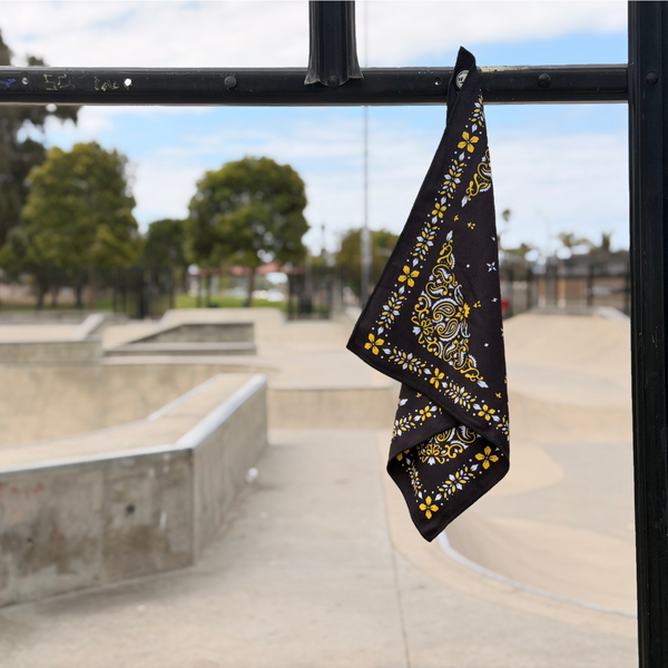 Happy Faced Bandito Skateboarding Towel magnetically attached to a metal skatepark fence. The microfiber skate towel showcases a black bandana-inspired design with intricate gold filigree, white floral accents, and traditional Western-style detailing. The built-in magnet keeps the towel securely attached between skate sessions.