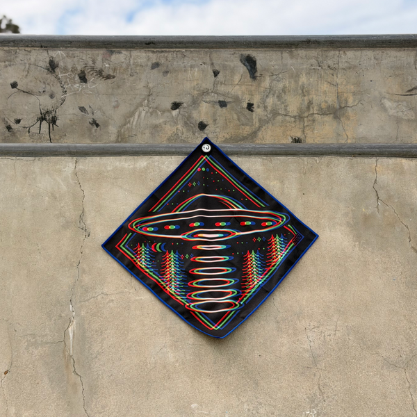 Happy Faced Beam Rider Skateboarding Towel displayed on a concrete skatepark ledge. The artist-designed microfiber skate towel features a retro UFO hovering above a forest landscape with colorful red, blue, and green offset graphics, celestial details, and a glowing tractor beam creating a vintage 3D effect.
