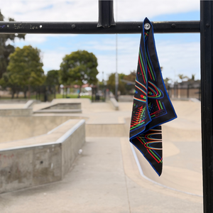Happy Faced Beam Rider Skateboarding Towel magnetically attached to a metal skatepark fence. The microfiber skate towel showcases a UFO flying over a pine forest with vibrant red, blue, and green retro 3D-style artwork, stars, moon phases, and a glowing tractor beam. The built-in magnet keeps the towel securely attached while skating.