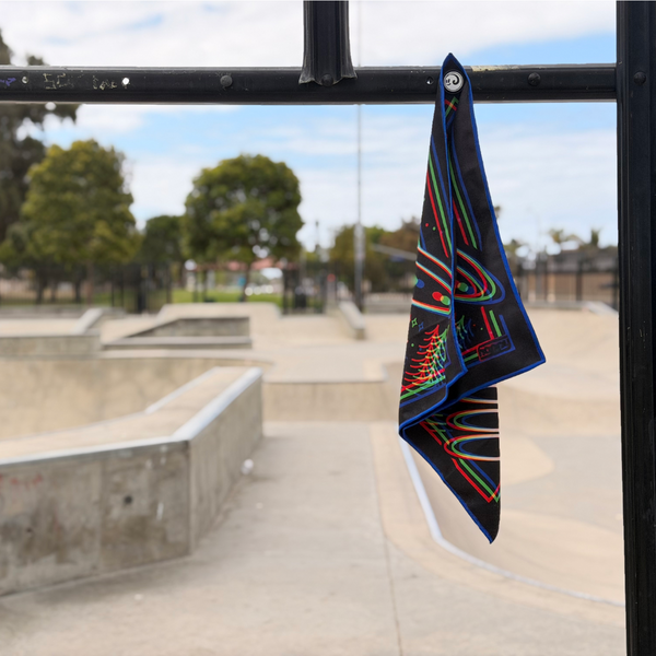 Happy Faced Beam Rider Skateboarding Towel magnetically attached to a metal skatepark fence. The microfiber skate towel showcases a UFO flying over a pine forest with vibrant red, blue, and green retro 3D-style artwork, stars, moon phases, and a glowing tractor beam. The built-in magnet keeps the towel securely attached while skating.