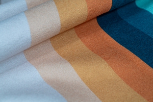 A close-up of a folded towel showcasing a series of horizontal stripes in soft, warm tones. The stripes range from light peach and beige to burnt orange and deep teal, creating a smooth gradient. The fabric appears soft and plush, with fine details in the texture, emphasizing the high quality and craftsmanship of the towel. The overlapping folds add depth and showcase the vibrant colors and clean design.