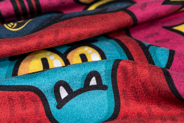 A close-up of a towel featuring a playful, colorful design with cartoon-like characters. The visible section shows part of a blue creature with small white fangs and large, yellow eyes. The towel has vibrant red, pink, and teal colors, with bold black outlines adding definition to the character. The fabric appears soft and textured, with the design showing a fun, whimsical style that brings a cheerful and lighthearted vibe to the towel.Happy Faced towel Creatures Comfort designed by Wotto