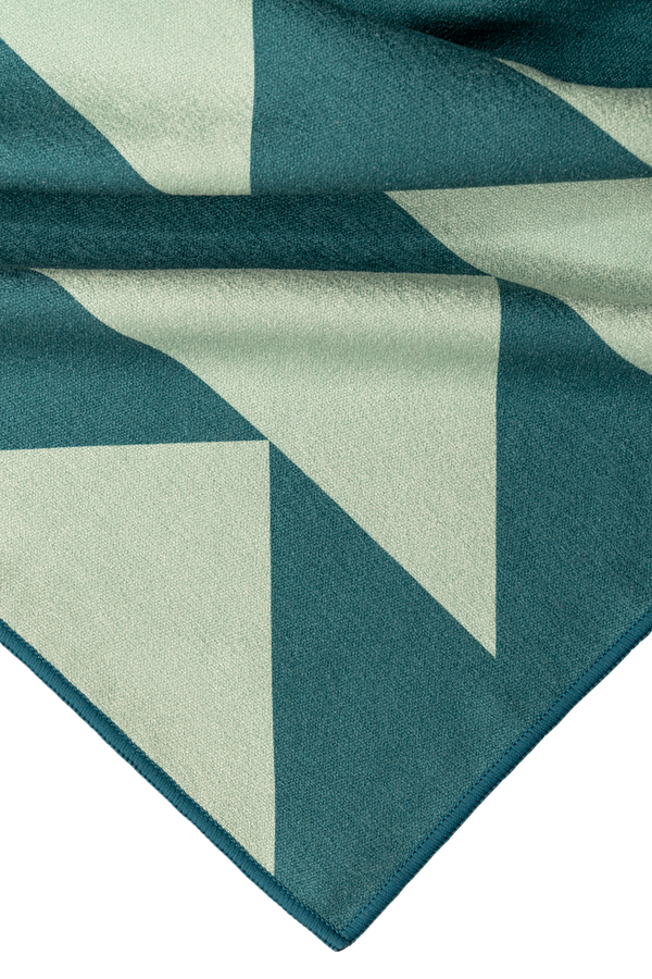 A close-up of a folded corner of a towel featuring a geometric design with teal and light green triangular shapes. The fabric's texture appears soft and smooth, with neatly stitched edges in a matching teal color. The abstract pattern of overlapping triangles creates a modern, clean look. The high-quality craftsmanship and bold design elements emphasize the towel's durability and style.