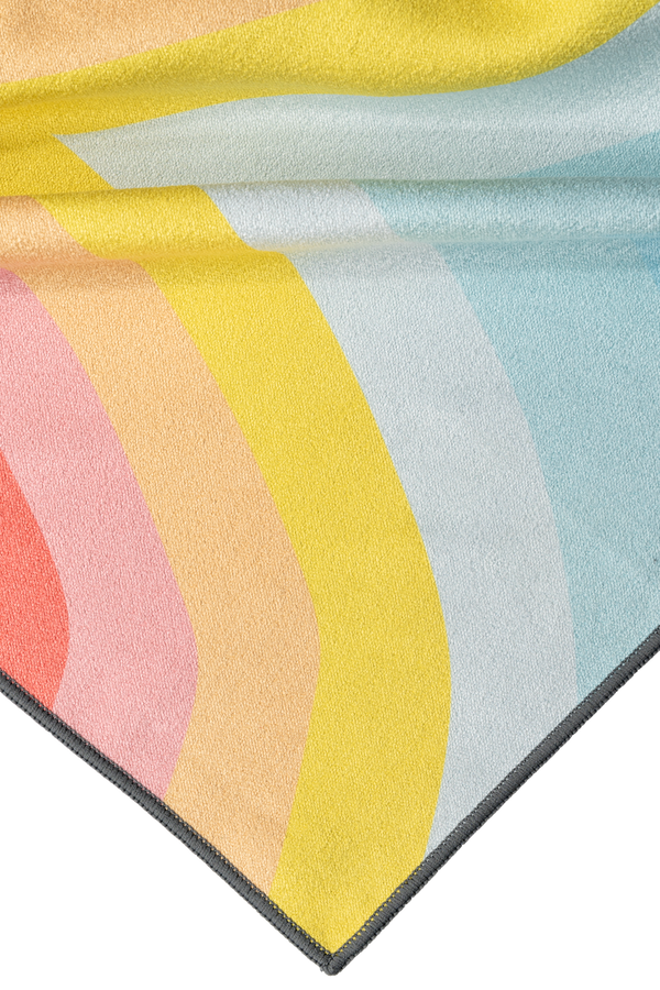 A close-up of a towel corner featuring a vibrant gradient design with flowing layers of yellow, pink, orange, and light blue hues. The soft fabric folds showcase the smooth transition between the colors, creating a playful and artistic look. The edge is neatly finished with dark stitching, highlighting the high-quality craftsmanship of the towel.