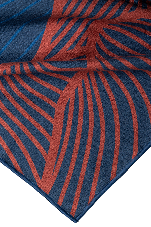 A close-up of a folded corner of a towel featuring a detailed abstract design. The towel is primarily dark blue with rust-red wavy lines that create a flowing, organic pattern. The fabric's texture is soft and smooth, with the stitching along the edge visible. The intricate lines and rich colors give the towel a bold and artistic appearance, emphasizing quality and craftsmanship.