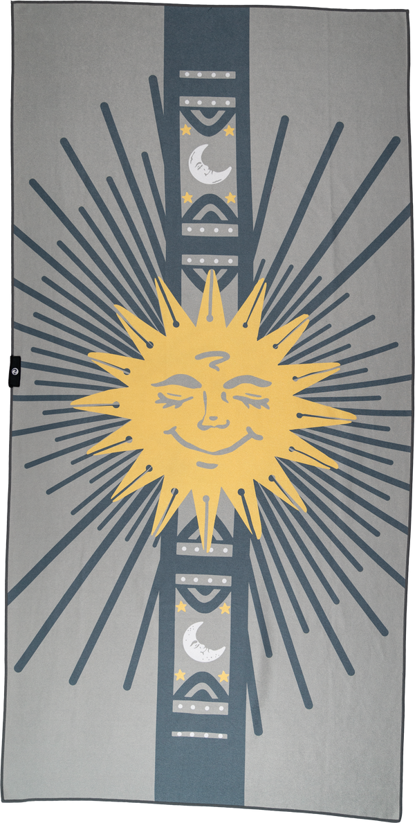 A towel featuring a large, smiling yellow sun at its center, with radiating rays in dark gray extending outward. A vertical band runs through the center with crescent moons and stars framed by geometric patterns in gray and teal. The overall design is symmetrical, combining celestial elements with bold, graphic details. The contrast of the bright sun against the gray background creates a playful yet serene cosmic-inspired aesthetic.