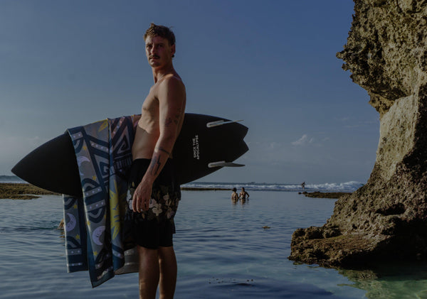 A Happy Faced Towel called Cosmic Currents draped over a surfboard held by a man beside the ocean