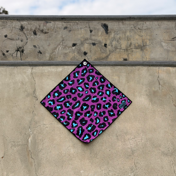 Happy Faced Disco Cheeta Skateboarding Towel displayed on a concrete skatepark ledge. The artist-designed microfiber skate towel features a vibrant purple leopard print pattern with black spots and electric blue highlights, combining bold animal print aesthetics with playful retro skateboarding style.