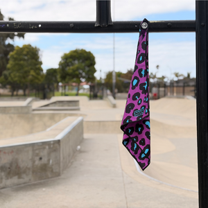 Happy Faced Disco Cheeta Skateboarding Towel magnetically attached to a metal skatepark fence. The microfiber skate towel showcases a striking purple leopard print pattern with black and electric blue accents inspired by retro fashion, punk graphics, and skateboarding culture. The built-in magnet keeps the towel securely attached and within easy reach during skate sessions.