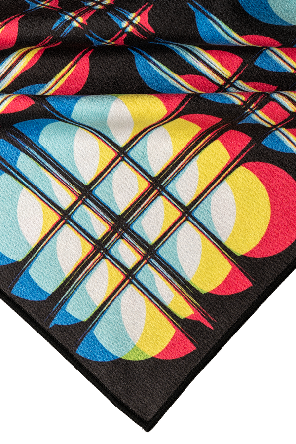 A close-up of a colorful geometric pattern featuring intersecting lines and bold shapes in vibrant reds, blues, yellows, and white. The design creates an eye-catching, plaid-like effect that adds depth and energy to the towel.