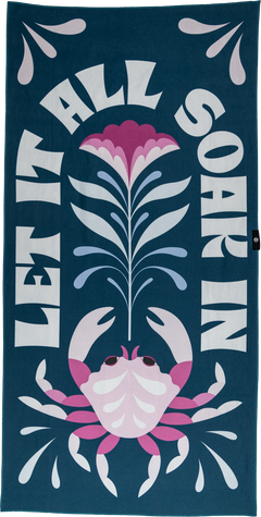 A playful towel with a pink crab, flower, and water droplets, framed by the bold text "LET IT ALL SOAK IN." The dark teal background and bright accents create a fun, vibrant beach vibe.