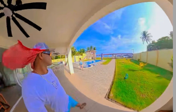 A man wearing a light blue t-shirt, a white cap, and a red mesh Happy Faced Cooling Towel stands under a covered patio area looking out toward a backyard with a swimming pool, green lawn, and blue sky.
