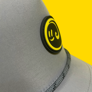 Close up view of the grey happy faced cap with a view of the rubber yellow and black logo on a yellow background
