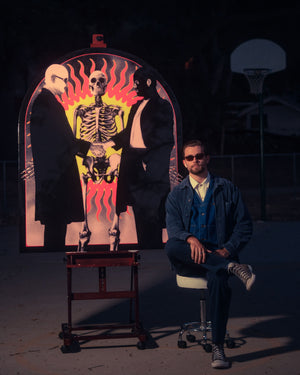 Photo realistic pop artist Hunter Blaze Pearson. Man in sunglasses wearing denim and converse sitting in front of a painting of a skeleton.