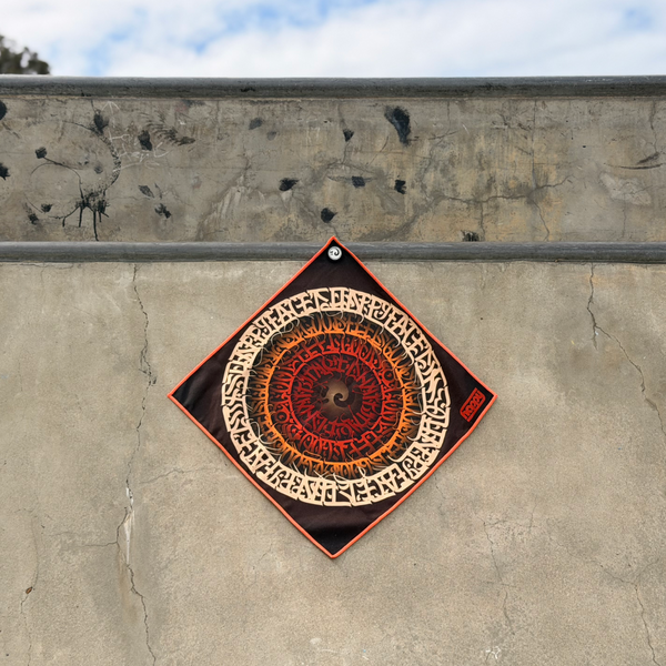 Happy Faced It's All Gravy Skateboarding Towel displayed on a concrete skatepark ledge. The artist-designed microfiber skate towel features concentric rings of hand-drawn lettering spiraling toward a central vortex in shades of cream, orange, and deep red, creating a bold psychedelic graphic inspired by skateboarding, street art, and vintage poster design.