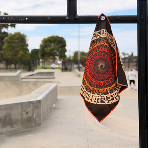 Happy Faced It's All Gravy Skateboarding Towel magnetically attached to a metal skatepark fence. The microfiber skate towel showcases a hypnotic spiral of hand-drawn lettering radiating inward through warm cream, orange, and red tones around a central vortex. The built-in magnet keeps the towel securely attached and easy to access during skate sessions.