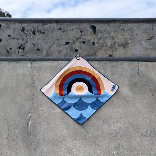 Happy Faced Rainbow Coast Skateboarding Towel displayed on a skatepark ledge. The microfiber skate towel features a vintage rainbow over stylized ocean waves in shades of blue, combining skateboarding, surf, and coastal-inspired artwork in a bold retro design.