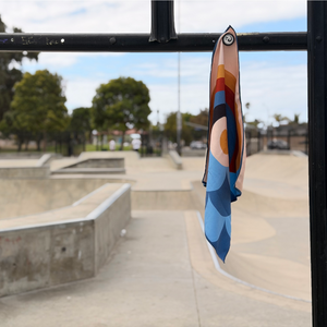 Happy Faced Rainbow Coast Skateboarding Towel hanging from a rail at a concrete skatepark. The artist-designed microfiber skate towel features a retro rainbow in warm earth tones above layered blue ocean waves, inspired by surf culture and the California coast.