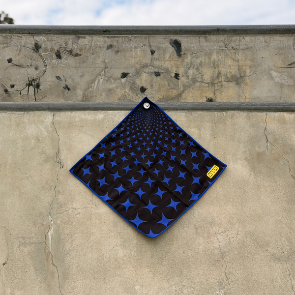 Happy Faced Super Star Skateboarding Towel displayed on a concrete skatepark ledge. The artist-designed microfiber skate towel features a repeating pattern of blue starbursts and black circular shapes that create a striking optical illusion effect, blending retro graphics with modern skateboarding style.