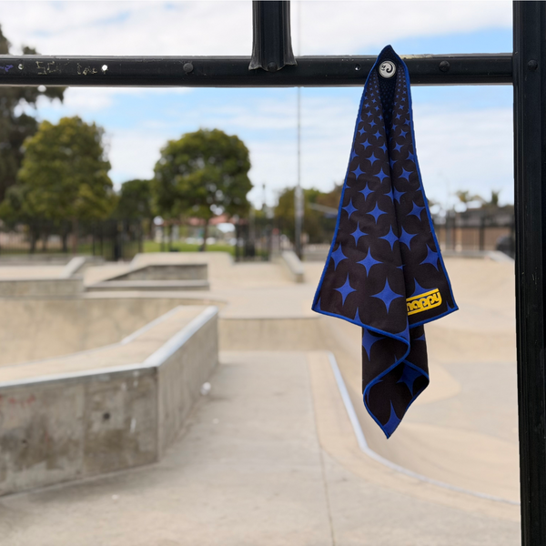 Happy Faced Super Star Skateboarding Towel magnetically attached to a metal skatepark fence. The microfiber skate towel showcases a bold blue and black geometric starburst pattern that creates depth and movement through a repeating optical illusion design. The built-in magnet keeps the towel securely attached and easy to access during skate sessions.