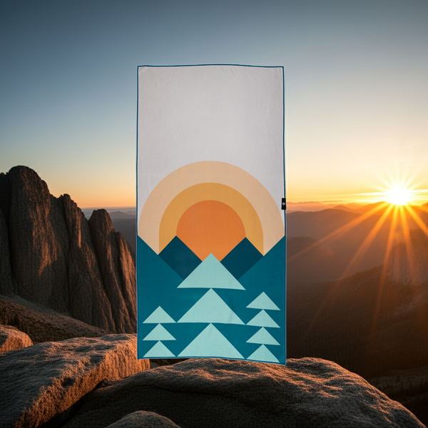 Towel with mountain and sun design held up against a mountainous landscape with sunset.