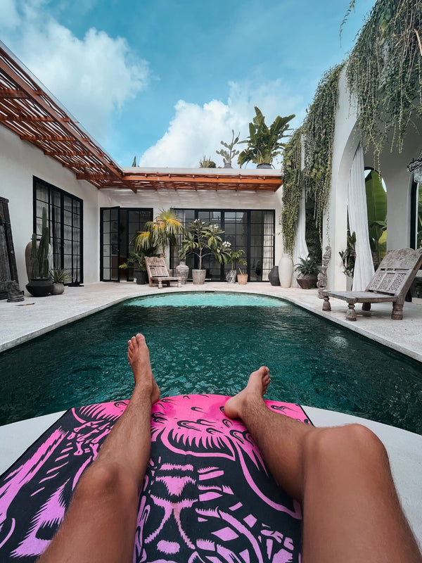 A person is relaxing by a pool, their legs stretched out on a vibrant pink and black Happy Faced towel with an intricate design. The setting is a stylish, modern villa with lush greenery, large glass windows, and potted plants scattered around the patio. Wooden furniture and plants add to the serene, tropical atmosphere. The clear, turquoise pool reflects the bright blue sky, which is partly covered by fluffy white clouds. The scene evokes a sense of calm and luxury in a private, tropical retreat.