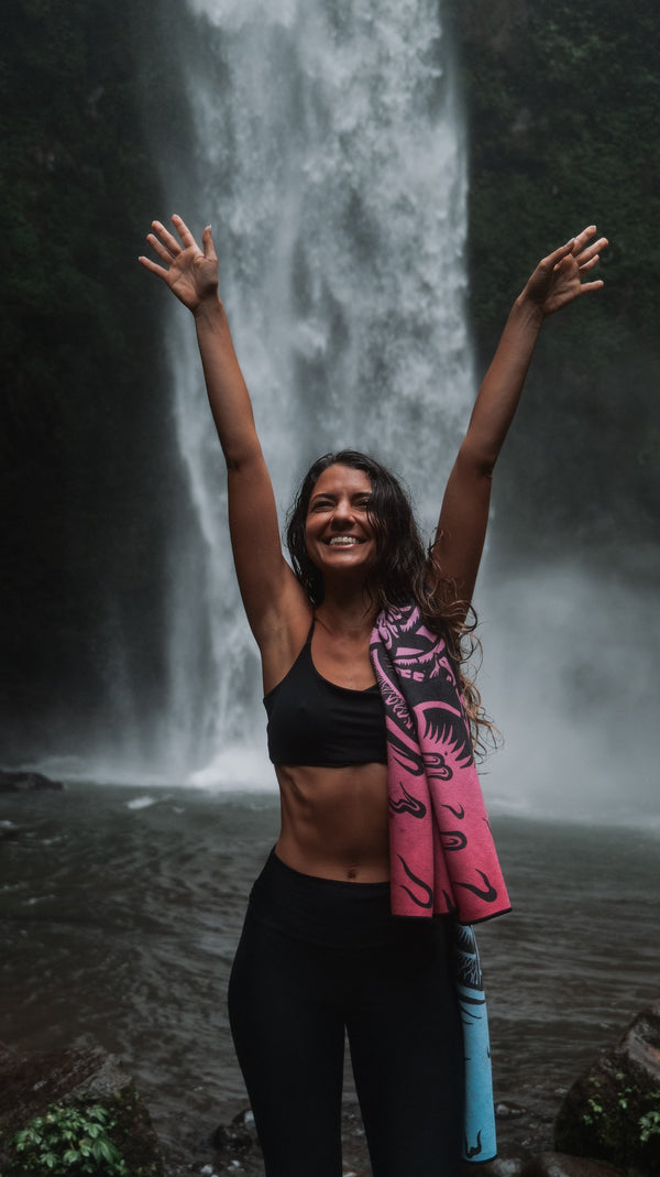 A woman stands in front of a powerful waterfall, smiling with her arms raised joyfully in the air. She is wearing a black sports bra and leggings, with a vibrant pink and blue towel draped over her shoulder. The mist from the waterfall surrounds her, and the lush greenery behind the falls adds to the sense of adventure and connection with nature. Her expression and body language exude happiness and freedom, capturing a moment of pure bliss in the wild outdoors.