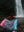 A woman stands at the base of a majestic waterfall, holding a vibrant towel with a gradient design from pink to blue, featuring intricate patterns. Her back is to the camera as she spreads the towel behind her, showcasing the artwork against the backdrop of lush greenery and cascading water. The rocky ground beneath her adds to the rugged, natural beauty of the scene. Her hair is damp, and she is dressed in athletic wear, fully immersed in the powerful and serene atmosphere of nature.