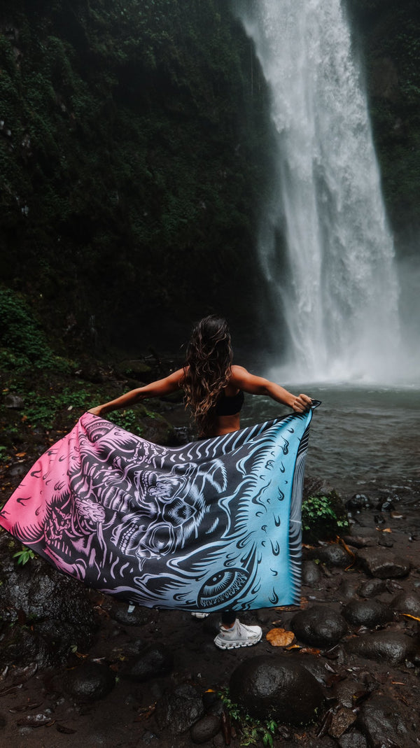 A woman stands at the base of a majestic waterfall, holding a vibrant towel with a gradient design from pink to blue, featuring intricate patterns. Her back is to the camera as she spreads the towel behind her, showcasing the artwork against the backdrop of lush greenery and cascading water. The rocky ground beneath her adds to the rugged, natural beauty of the scene. Her hair is damp, and she is dressed in athletic wear, fully immersed in the powerful and serene atmosphere of nature.