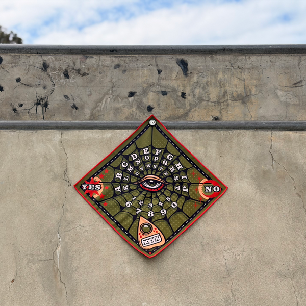Happy Faced Web Ritual Skateboarding Towel displayed on a skatepark ledge. The artist-designed microfiber skate towel features a vintage spirit board inspired graphic with a central eye, alphabet letters, numbers, mystical symbols, and a planchette set against a green spiderweb pattern with red accents.