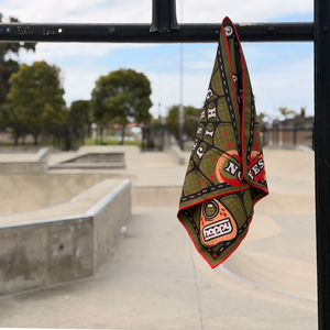 Happy Faced Web Ritual Skateboarding Towel magnetically attached to a fence at a skatepark. The artist-designed microfiber skate towel features a vintage spirit board graphic with a watchful eye, alphabet letters, numbers, a planchette, and spiderweb artwork accented with bold red trim, showcasing the towel's built-in magnet for easy storage while skating.