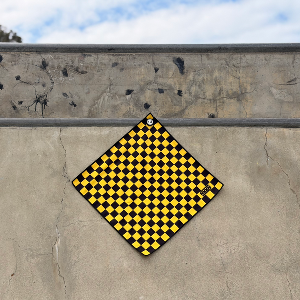 Happy Faced Yellow Checker Skateboarding Towel displayed on a concrete skatepark ledge. The artist-designed microfiber skate towel features a bold yellow and black checkerboard pattern inspired by classic skateboarding culture, combining a clean geometric design with timeless skate style.