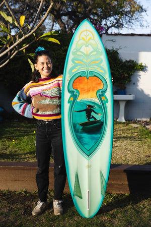 Hawaiian artist Angie Samblotte holding a turquoise surfboard she painted with a picture of a man surfing on it for Happy Faced Towels