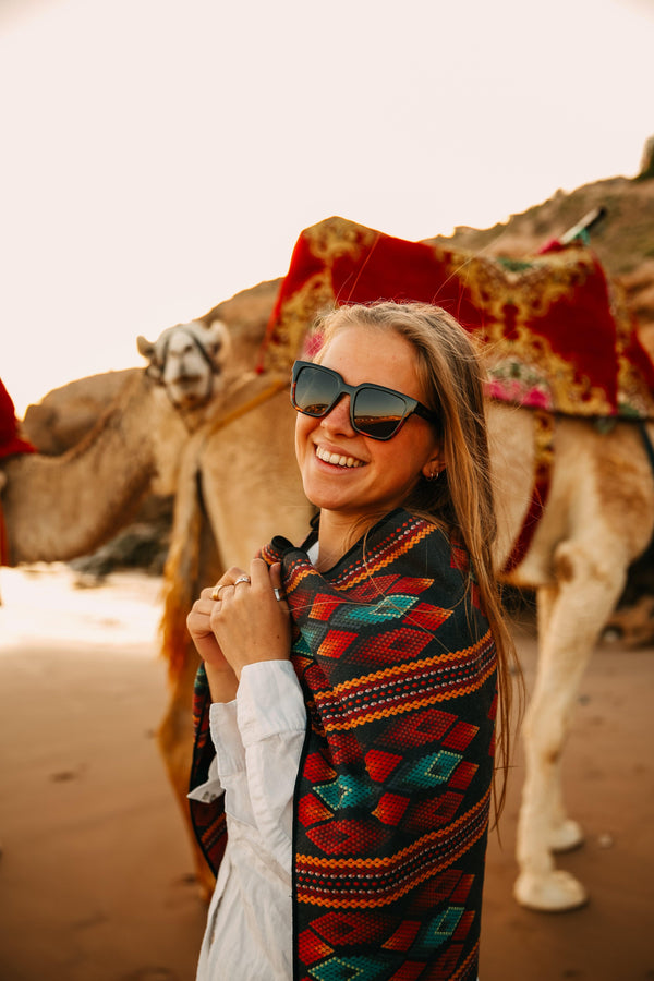 A woman smiling in Morocco with a small Happy Faced Towel over her shoulders. a squad of camels just hanging out in the background.