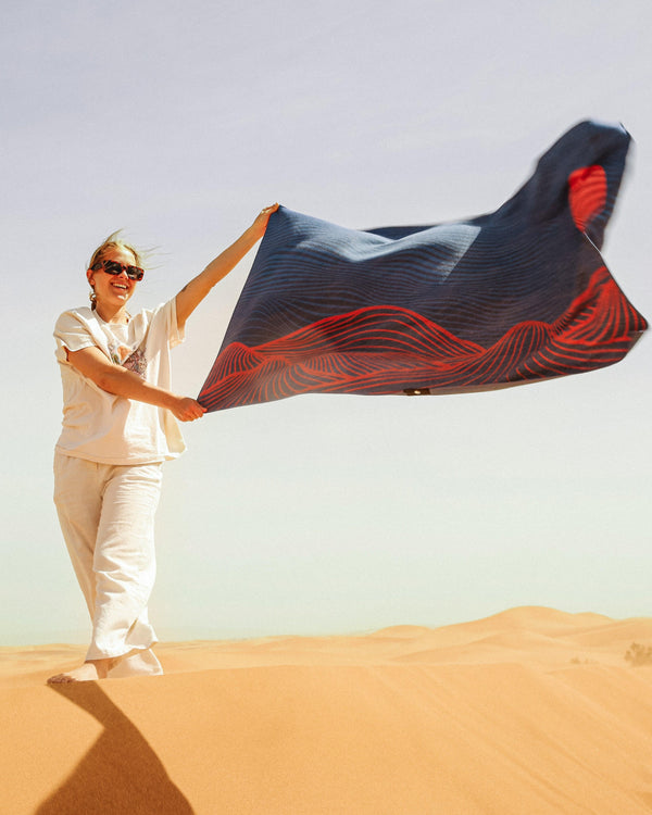 A smiling woman stands on a dune in the Sahara Desert beach holding a Happy Faced Towel that blows in the warm wind