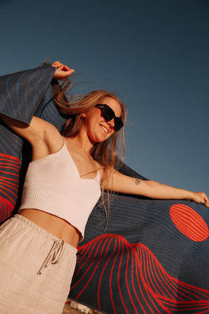 A smiling woman stands on the beach with a Happy Faced Towel draped over her shoulders and her arms are outstretched