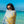 A woman wrapped in a towel with a bold sun design stands by the ocean, gazing at the camera while wearing round sunglasses. The towel features yellow and blue colors against a neutral background, adding a pop of brightness. The crystal-clear turquoise water and deep blue sky in the background create a perfect sunny beach day atmosphere. Her relaxed and playful expression, combined with the vibrant setting, evokes a carefree summer vibe.